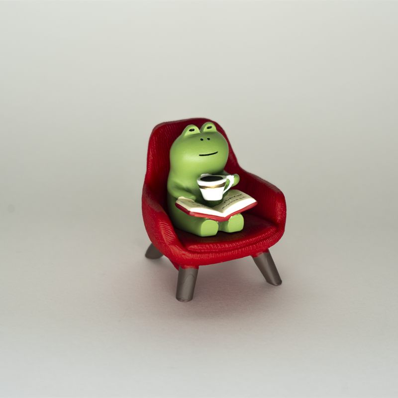 A small green frog figurine sitting in a red armchair with a book and a cup of coffee